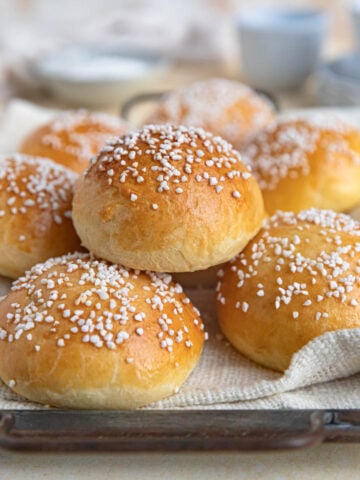 A stack of brioche au sucre (sugar brioche) buns on a cloth covered tray. The buns are topped Swedish pearl sugar. There is a tea cup and plates in the background