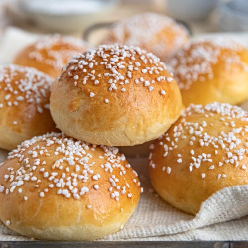 A stack of brioche au sucre (sugar brioche) buns on a cloth covered tray. The buns are topped Swedish pearl sugar. There is a tea cup and plates in the background