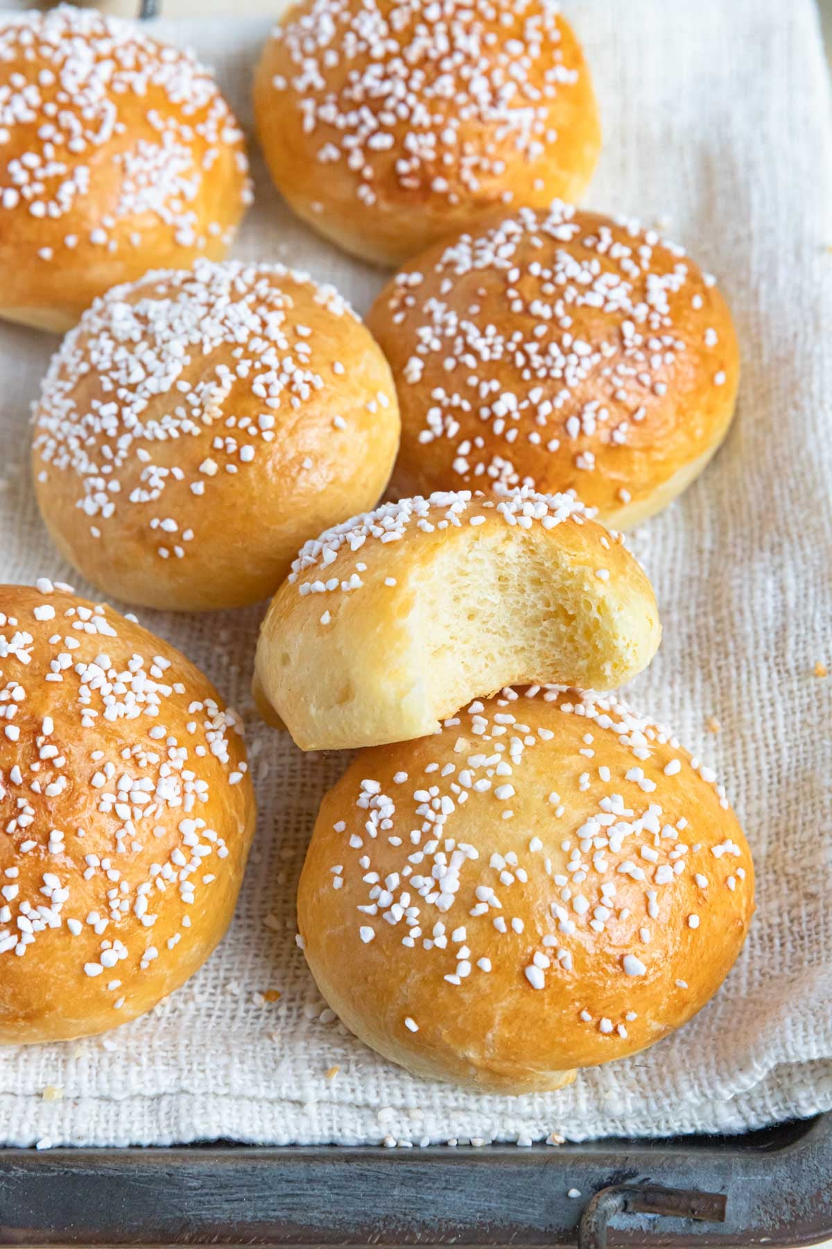 A stack of brioche au sucre (sugar brioche) buns on a cloth covered tray. The buns are topped Swedish pearl sugar. One of the buns has a bite taken out of it.