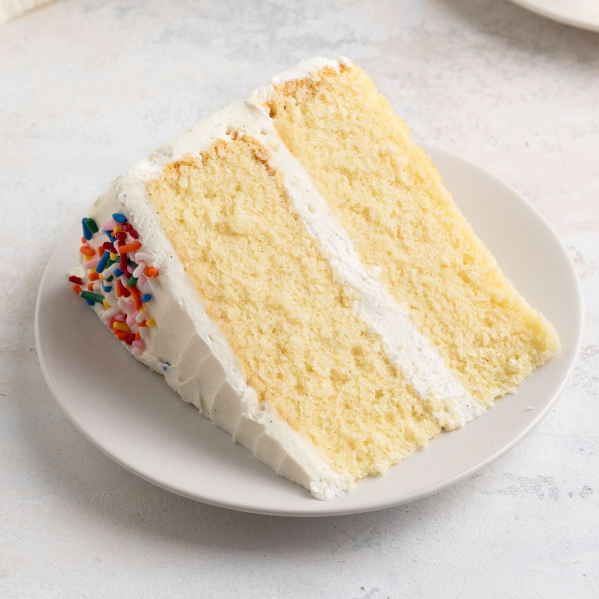A slice of vanilla cake on a dessert plate. The cake is two layers with vanilla buttercream frosting an decorated with sprinkles on top