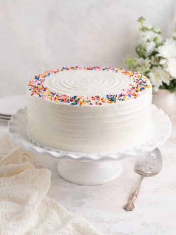 Vanilla layer cake with Swiss meringue buttercream on a ceramic cake stand. There is a silver plated server and a linen tea towel next to the cake stand.