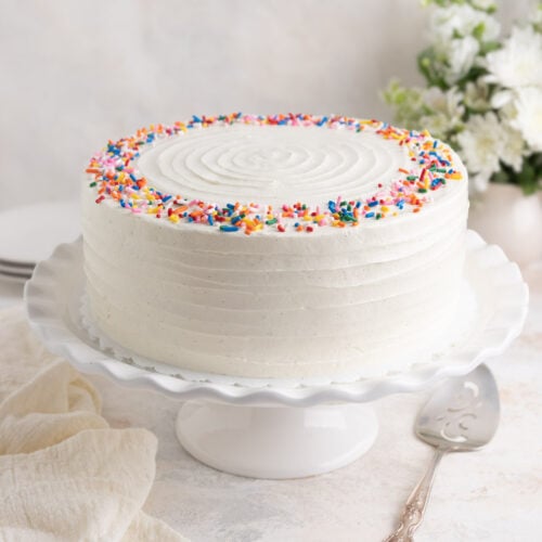 Vanilla layer cake with Swiss meringue buttercream on a ceramic cake stand. There is a silver plated server and a linen tea towel next to the cake stand.