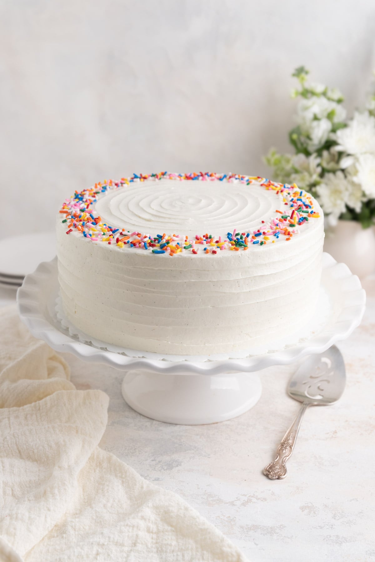 Vanilla layer cake with Swiss meringue buttercream on a ceramic cake stand. There is a silver plated server and a linen tea towel next to the cake stand.