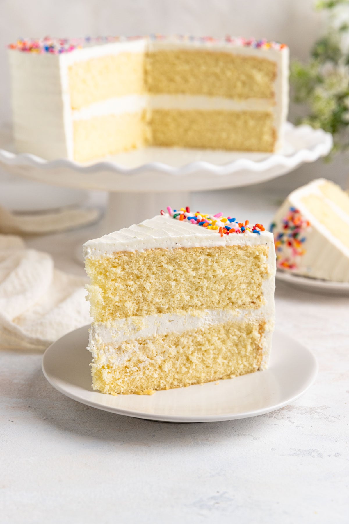 A slice of vanilla layer cake on a white dessert plate. The rest of the cake is in the background on a white cake stand.