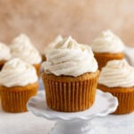A banana cupcake with vanilla buttercream on a mini cake stand. More frosted banana cupcakes are in the background
