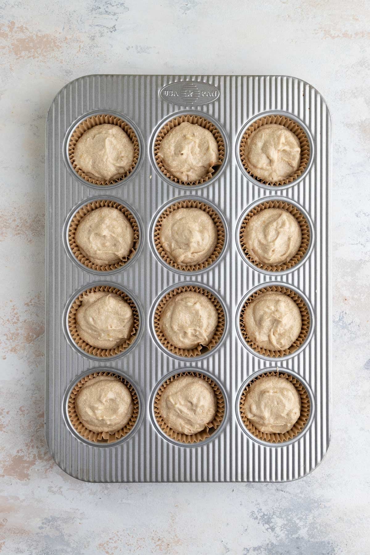 A 12-tin cupcake pan with brown liners and each liner filled with banana cupcake batter
