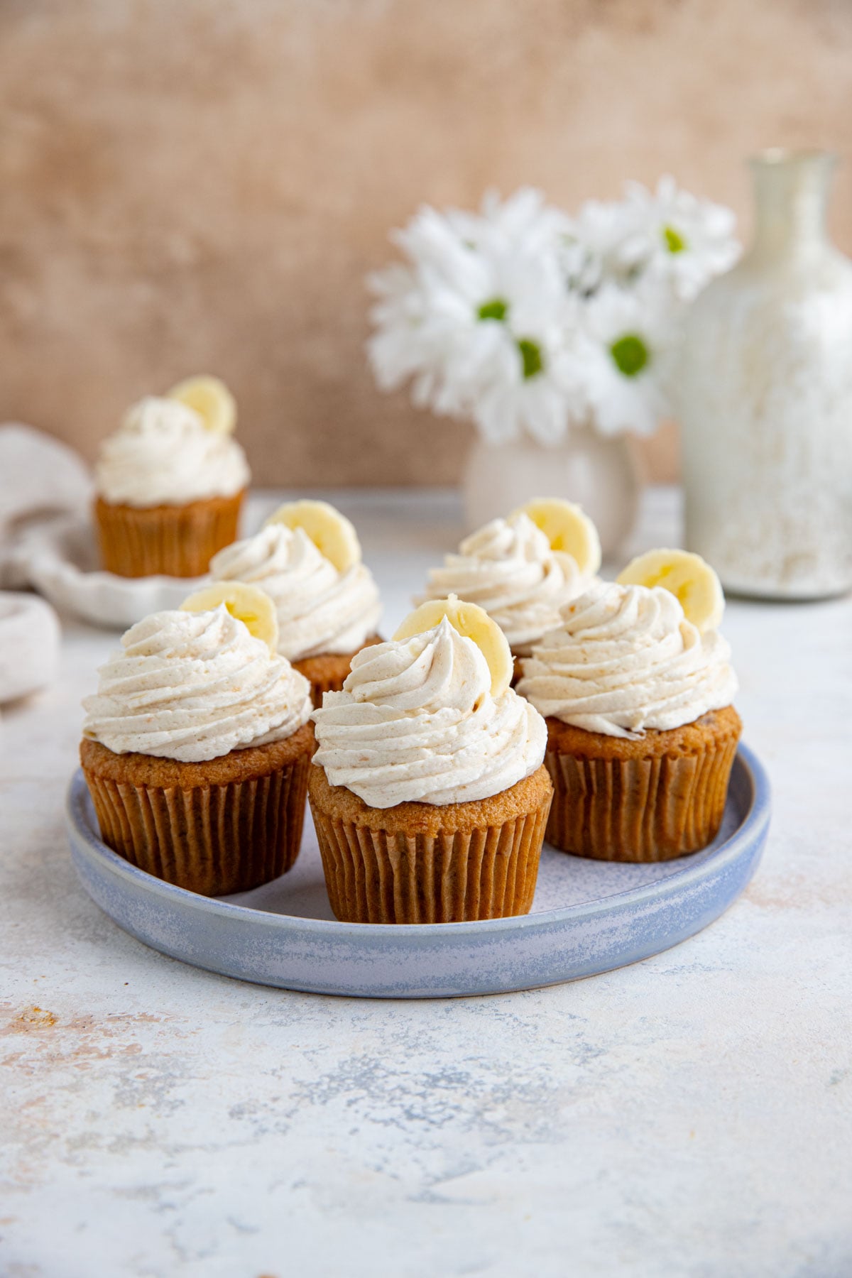 Five banana cupcakes with vanilla ermine frosting on a blue ceramic plate with one cupcake in the background on a small dessert plate. There is small vase of white flowers in the backgroud.