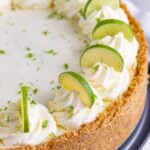 Key lime cheesecake with a graham cracker crust. The top of the cheesecake is decorated with piped whipped cream and half slices of key lime on top of the whipped cream