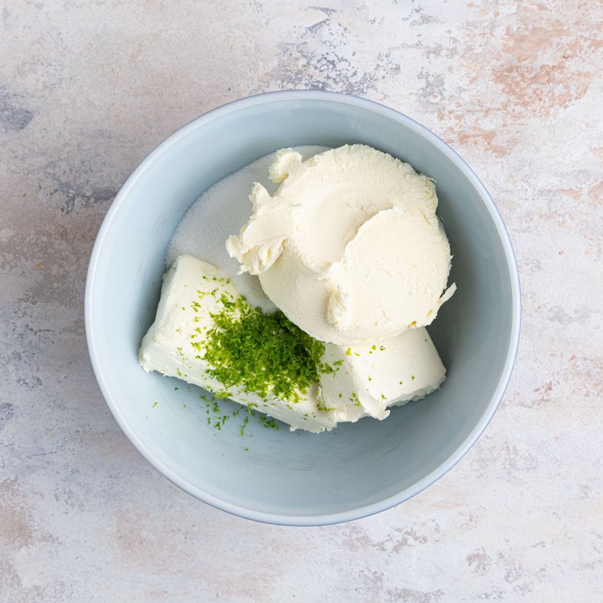 Lime zest, 16 ounces of cream cheese, sugar, and 8 ounces of mascarpone in a ceramic mixing bowl