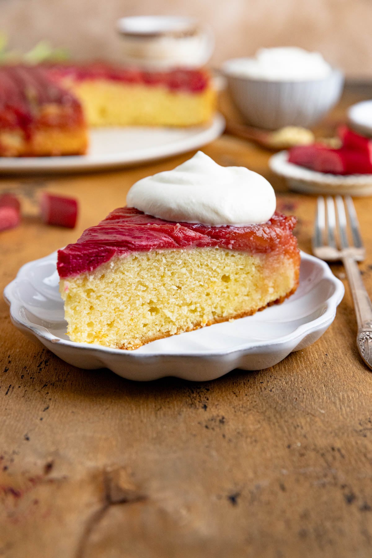 A slice of rhubarb upside down cake topped with a dollop of whipped cream on a small white plate. The rest of the cake is in the background. There is a fork next to the plate.