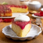 A slice of rhubarb upside down cake topped with a dollop of whipped cream on a small white plate. The rest of the cake and a bowl of whipped cream are in the background. There is a fork next to the plate.
