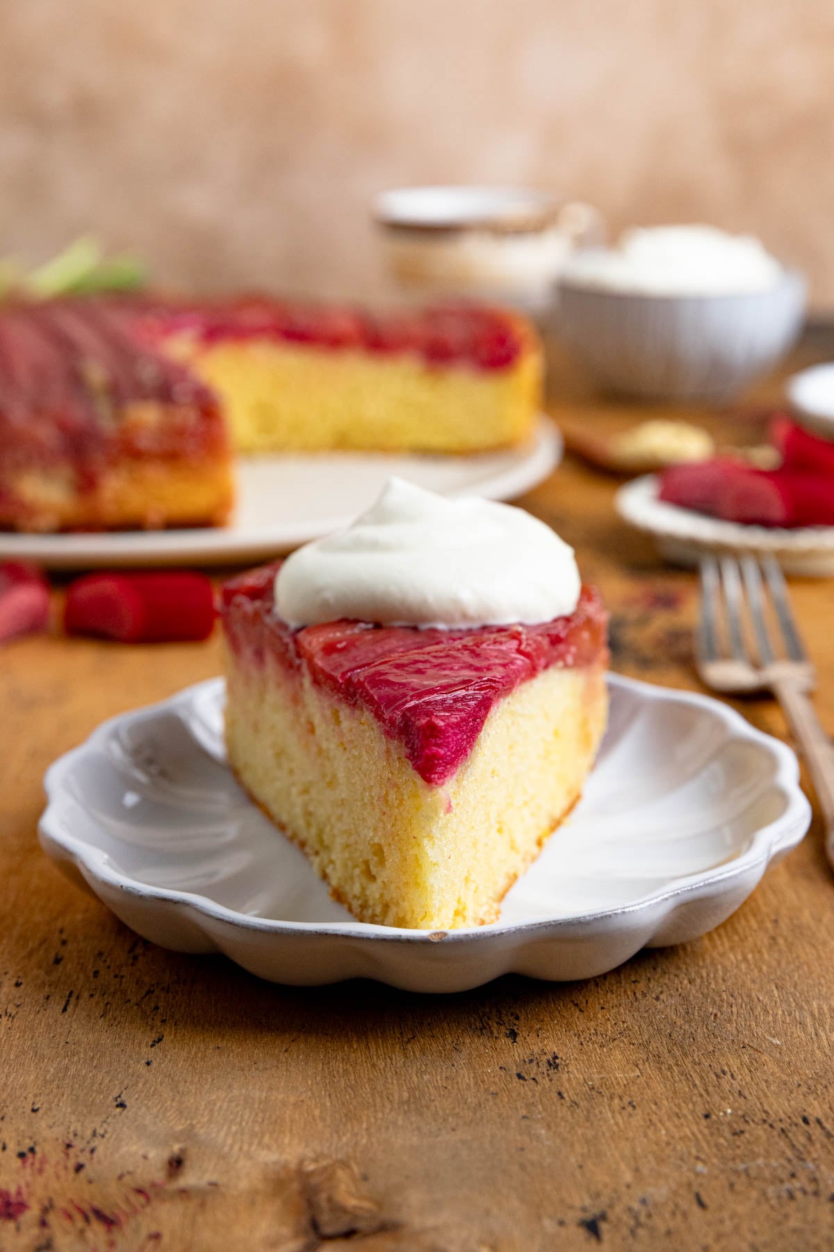 A slice of rhubarb upside down cake topped with a dollop of whipped cream on a small white plate. The rest of the cake and a bowl of whipped cream are in the background. There is a fork next to the plate.