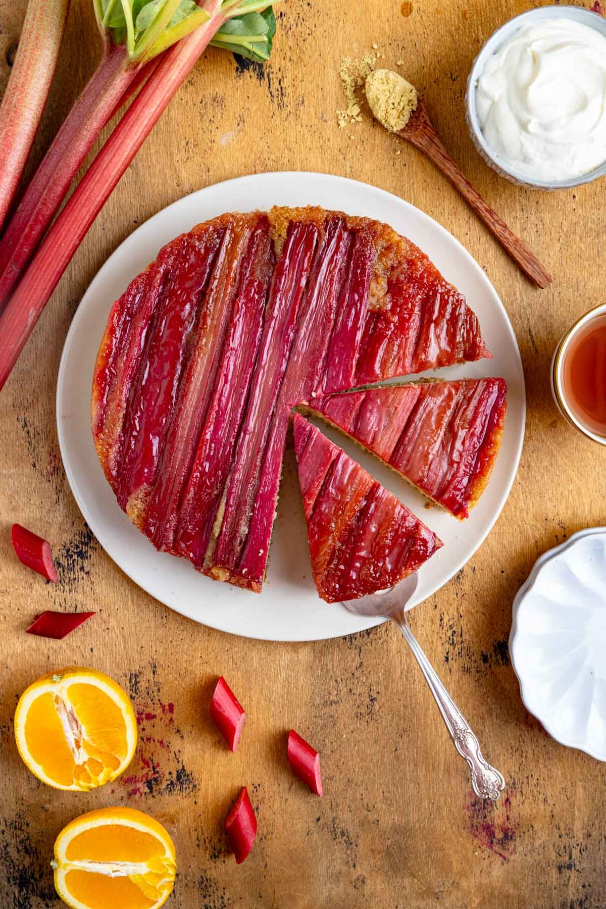 Rhubarb upside down cake on a white plate with two slices cut out and a cake server under one of the slices to lift the slice off the plate. There is a bowl of whipped cream and a cup of tea on the right side of the plate. There are stalks of rhubarb in the upper left corner and an orange cut in half in the lower left corner.