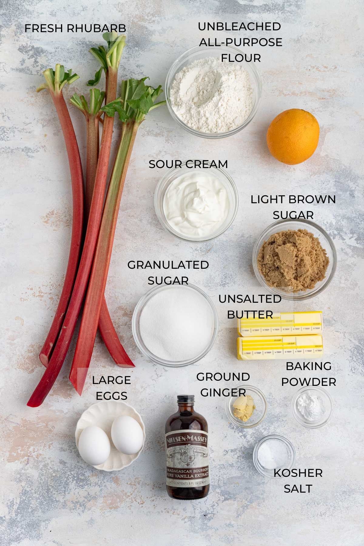 Rhubarb upside down cake ingredients arranged on a board. Clockwise - unbleached all-purpose flour, a fresh orange, sour cream, light brown sugar, unsalted butter, baking powder, kosher salt, ground ginger, granulated sugar, vanilla extract, large eggs, fresh rhubarb stalks