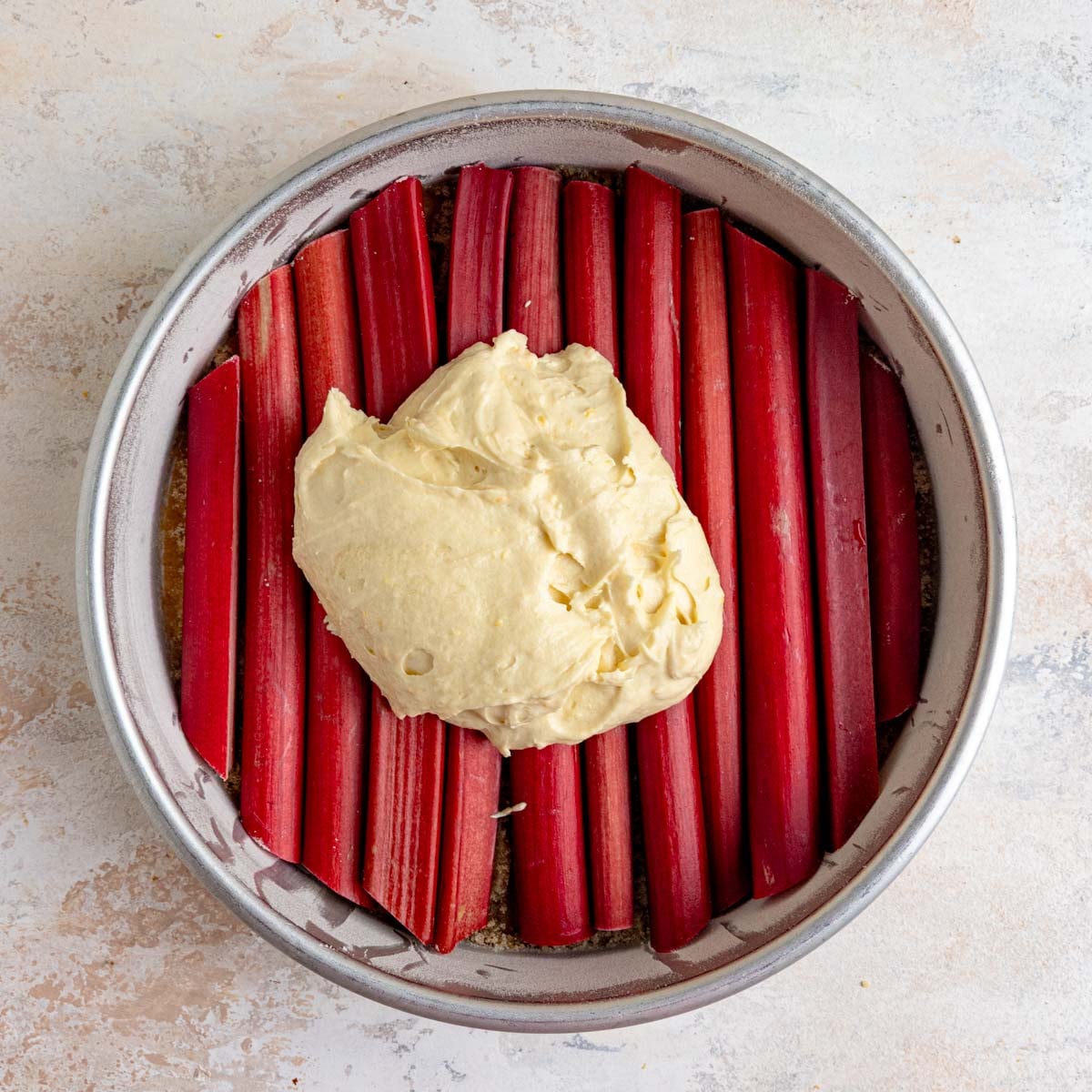 The round cake pan filled with red stalks of fresh rhubarb with a dollop of the batter on top of the rhubarb