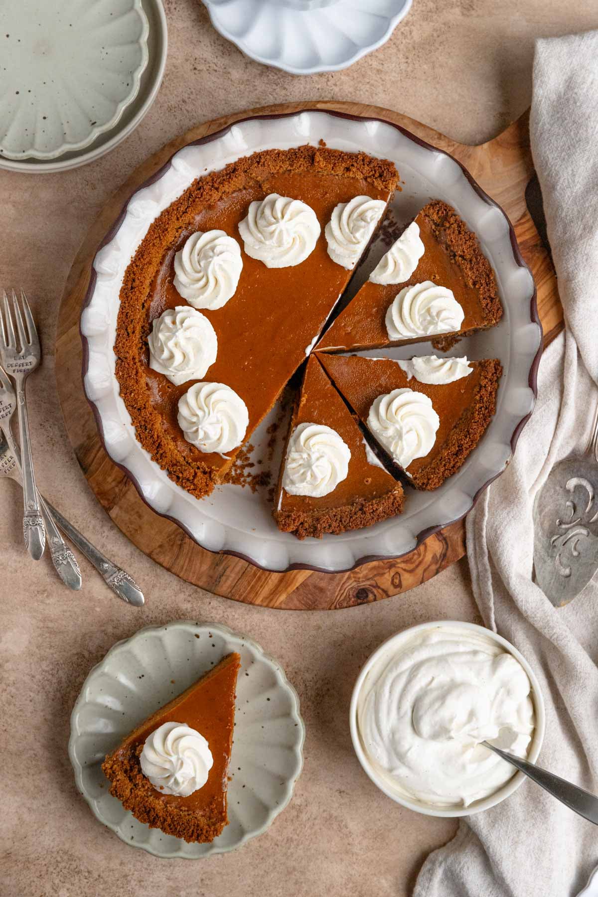 Sweet potato pie in a a ceramic pie dish. The top of the pie is decorated with swirls of whipped cream. There is a slice of pie on a dessert plate and a bowl of whipped cream next to the pie dish