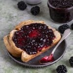 A slice of toast with a spread of blackberry jam on a small grey plate. An 8-ounce jar of blackberry jam next to the plate, and a small bowl of fresh blackberries next to the jar of jam.
