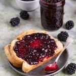 A slice of toast with a spread of blackberry jam on a small grey plate. An 8-ounce jar of blackberry jam next to the plate, and a small bowl of fresh blackberries next to the jar of jam.