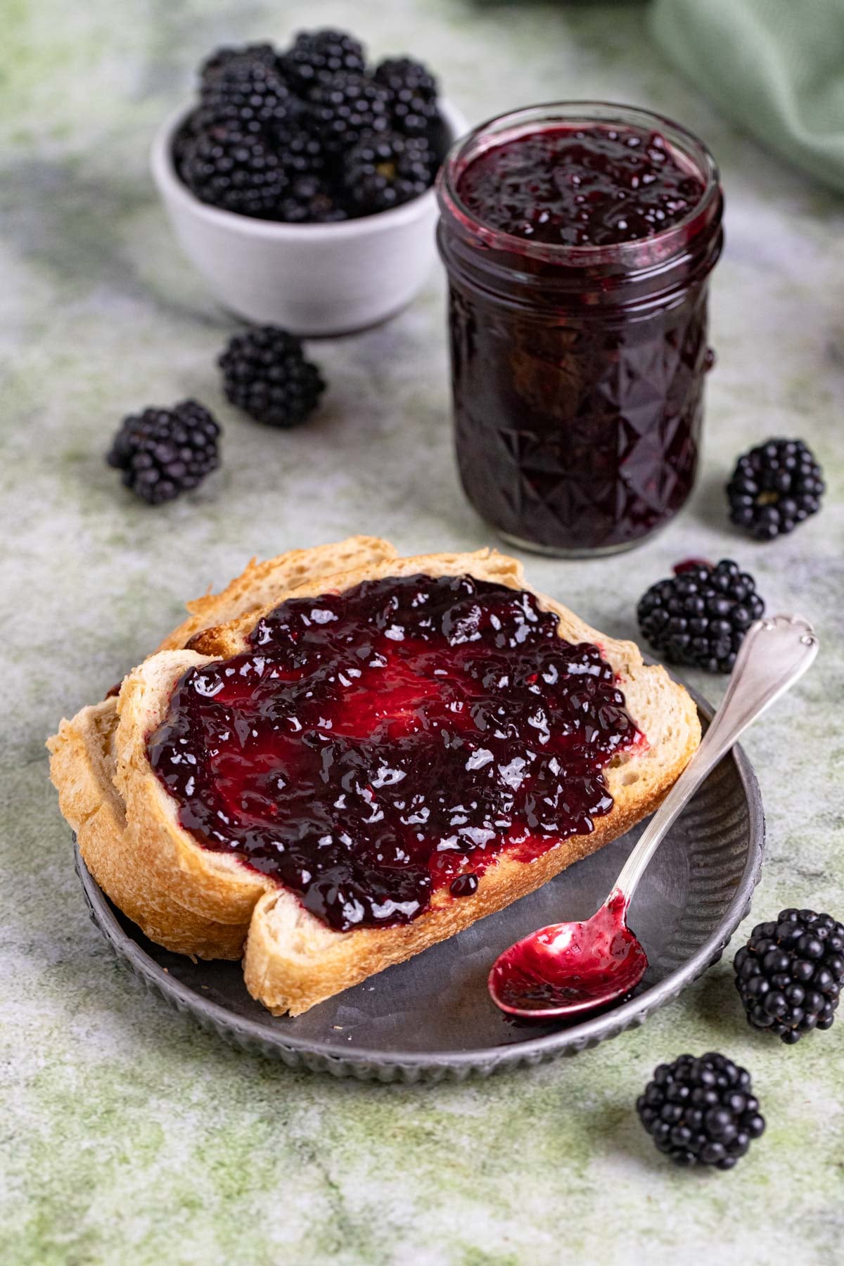 A slice of toast with a spread of blackberry jam on a small grey plate. An 8-ounce jar of blackberry jam next to the plate, and a small bowl of fresh blackberries next to the jar of jam.