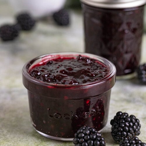 Two jars of blackberry jam on a table with a small bowl blackberries. One jar has a lid, the other doesn't