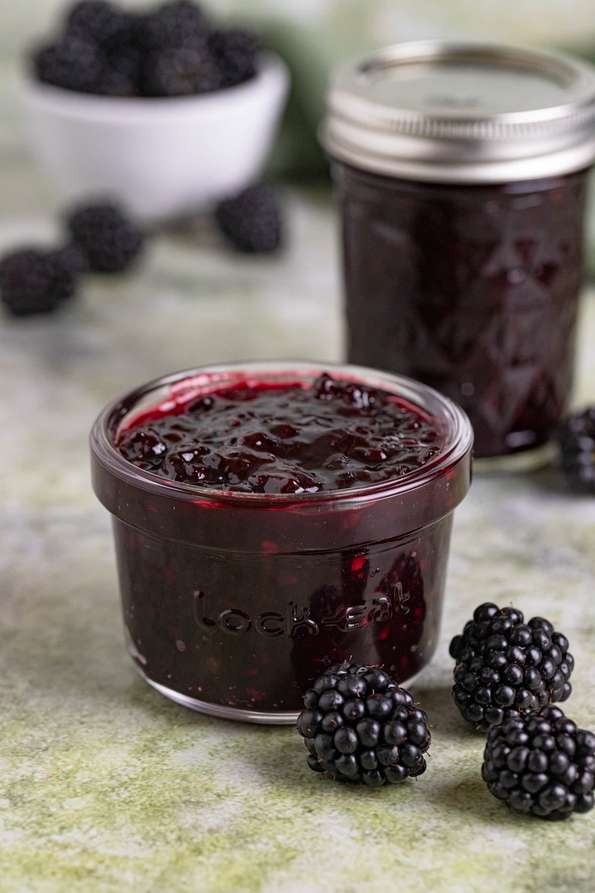 Two jars of blackberry jam on a table with a small bowl blackberries. One jar has a lid, the other doesn't