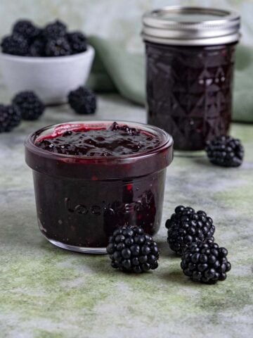 Two jars of blackberry jam on a table with a small bowl blackberries. One jar has a lid, the other doesn't