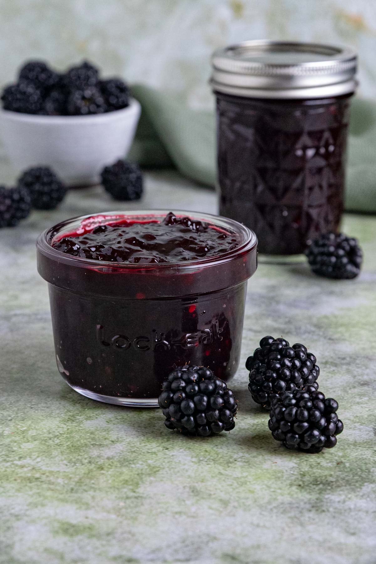 Two jars of blackberry jam on a table with a small bowl blackberries. One jar has a lid, the other doesn't