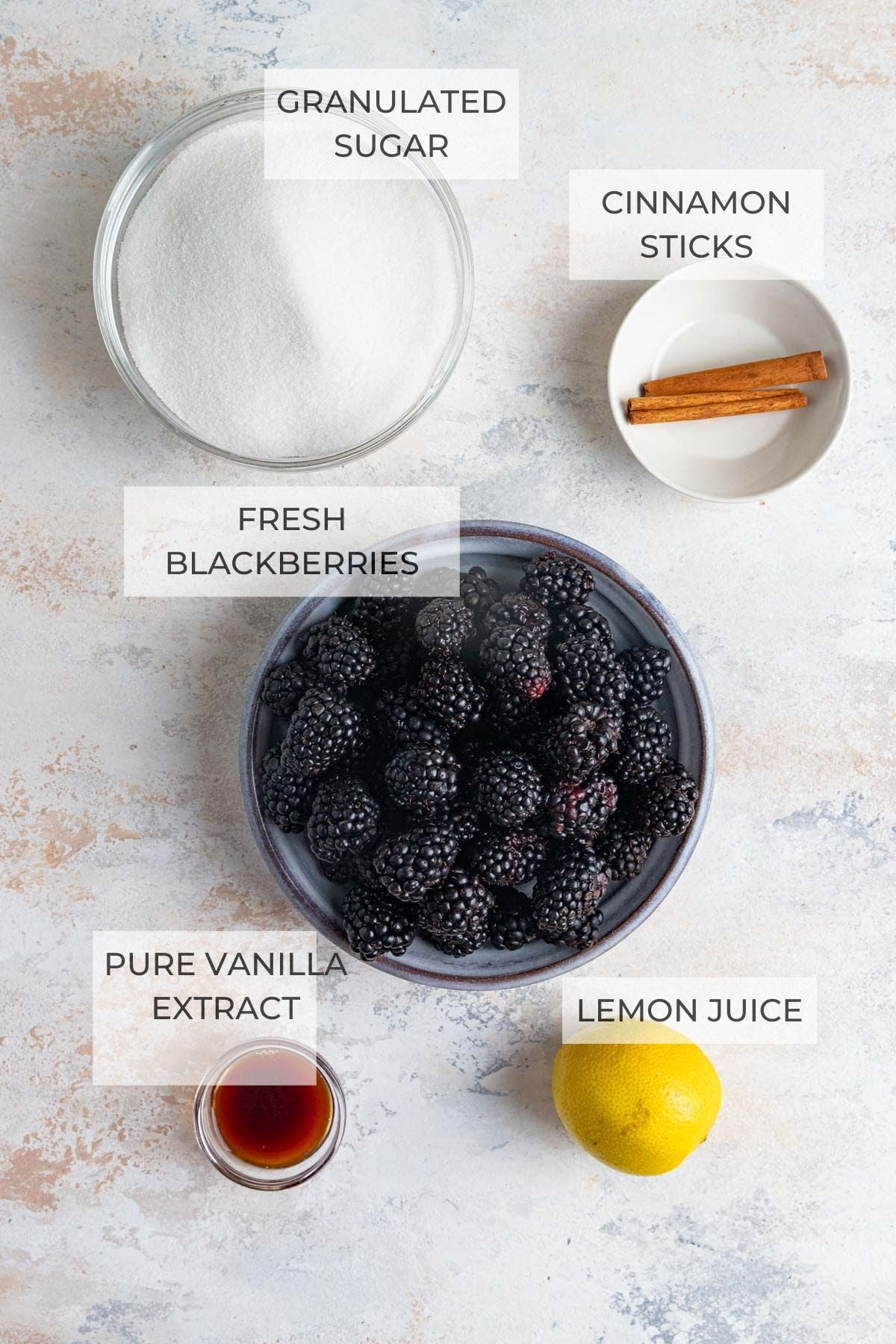 Blackberry jam ingredients on a flat surface: granulated sugar, cinnamon sticks, a lemon, pure vanilla extract, fresh blackberries
