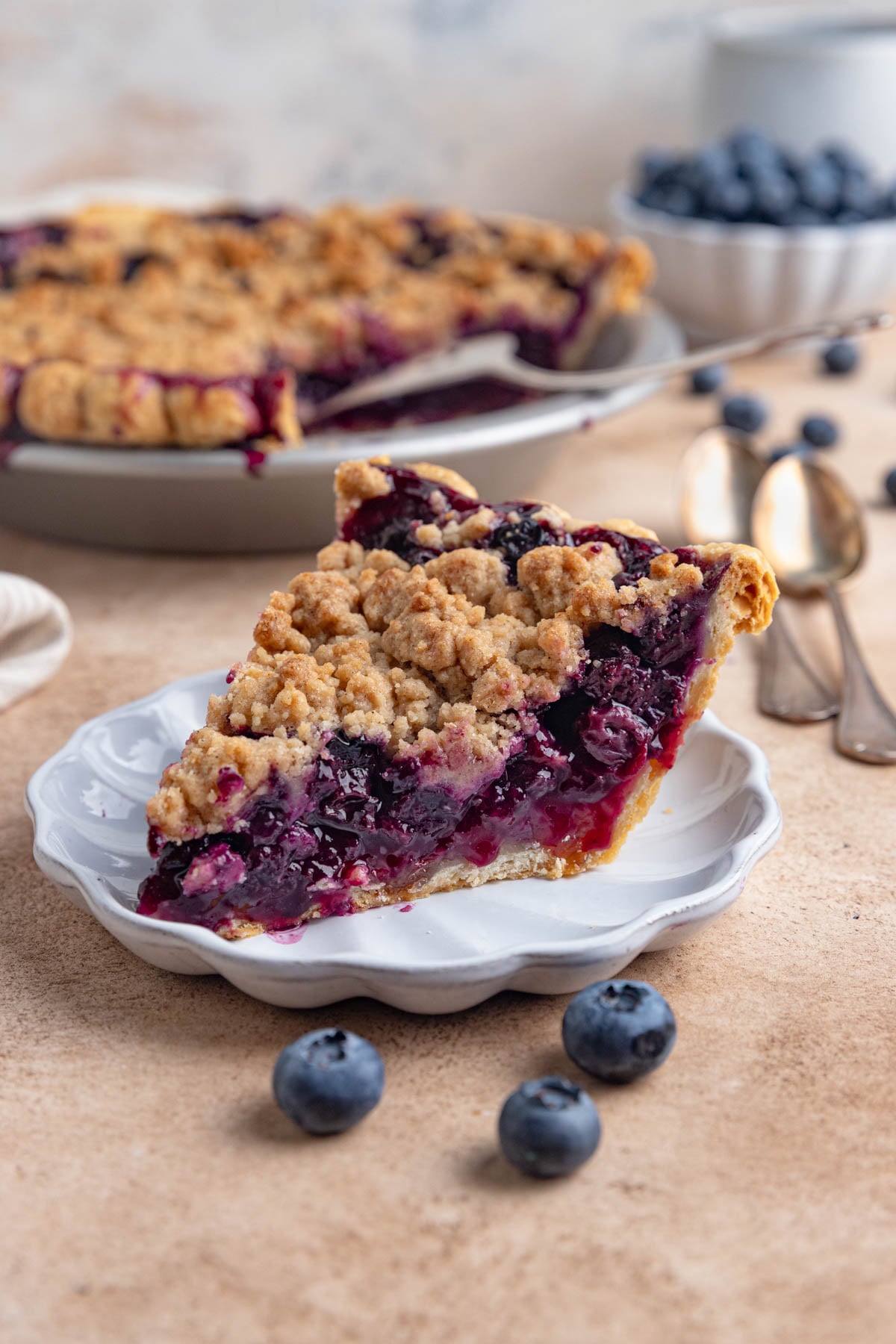 A slice of blueberry pie on a small white plate. The rest of pie in a pie plate is in the background.