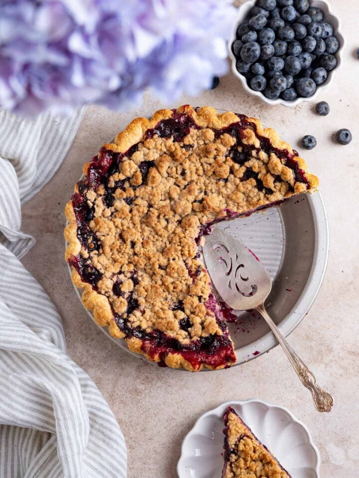 Blueberry crumble pie with two slices cut out. One slice on a small plate next to the pie and a pie server in the pie plate. A small bowl of fresh blueberries is next to the pie.