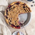 Blueberry crumble pie with two slices cut out. One slice on a small plate next to the pie and a pie server in the pie plate. A small bowl of fresh blueberries is next to the pie.