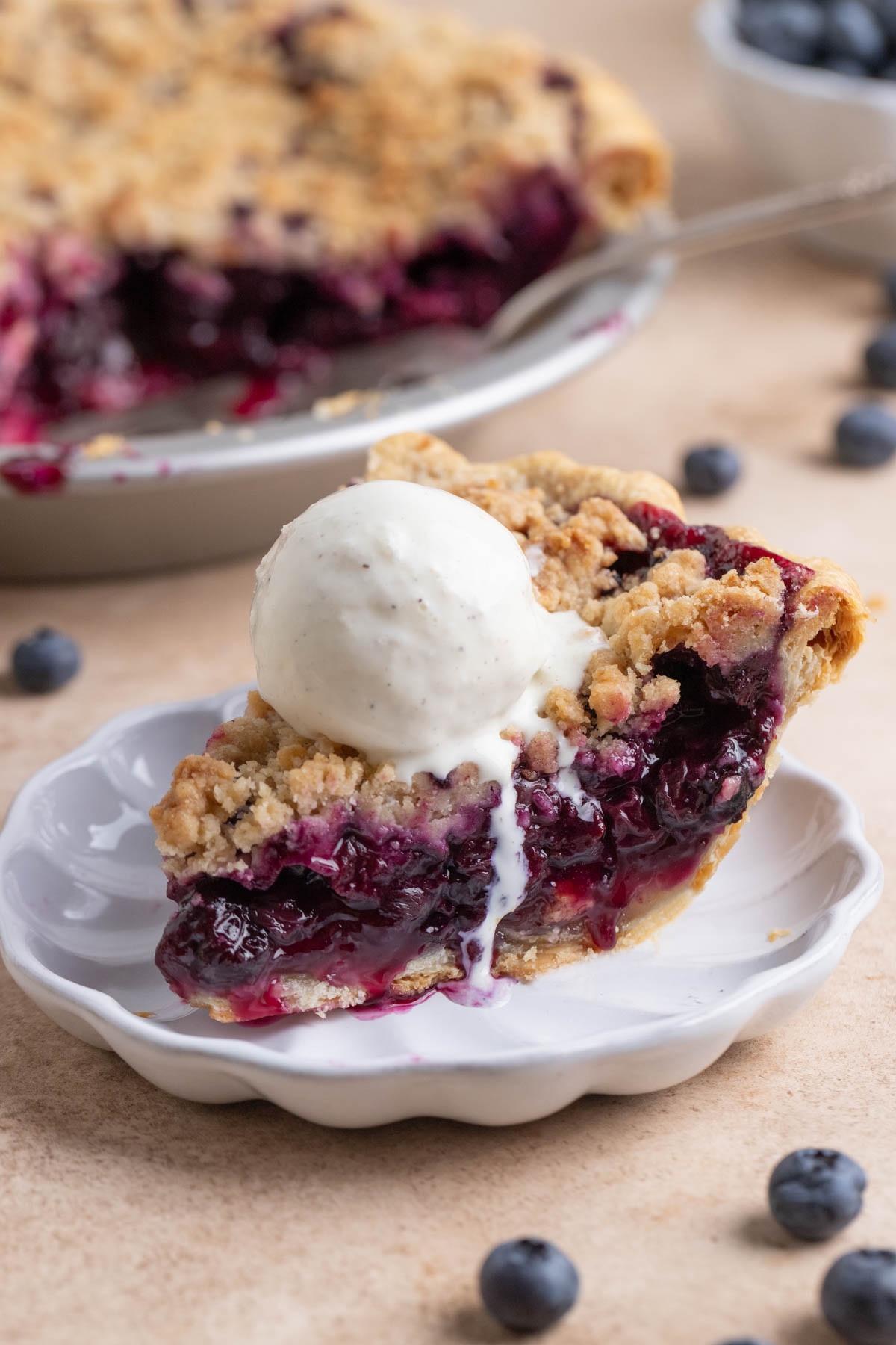 A slice of blueberry pie on a small white plate with a scoop of vanilla ice cream on top of the pie. The rest of pie in a pie plate is in the background.