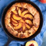 The baked peach galette in a cast-iron pan with two pieces cut out. One slice is on a small plate next to the cast iron pan and there are fresh peaches arranged next to the pan.