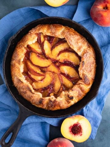 The baked peach galette in a cast-iron pan with two pieces cut out. One slice is on a small plate next to the cast iron pan and there are fresh peaches arranged next to the pan.