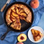 The baked peach galette in a cast-iron pan with two pieces cut out. One slice is on a small plate next to the cast iron pan and there are fresh peaches arranged next to the pan.