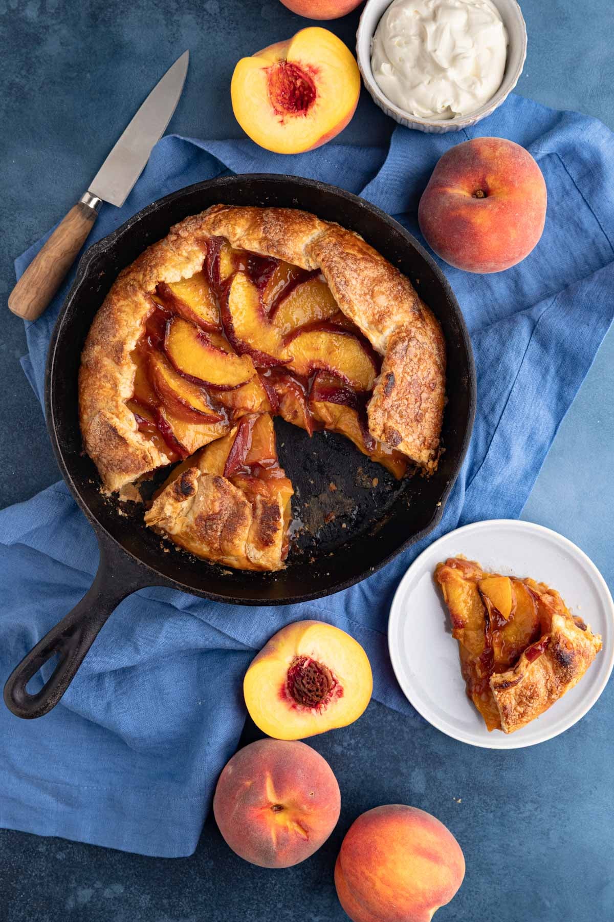 The baked peach galette in a cast-iron pan with two pieces cut out. One slice is on a small plate next to the cast iron pan and there are fresh peaches arranged next to the pan.