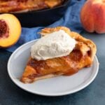 A slice of the peach galette with a dollop of whipped cream on a small plate with the rest of the peach galette and fresh peaches in the background.