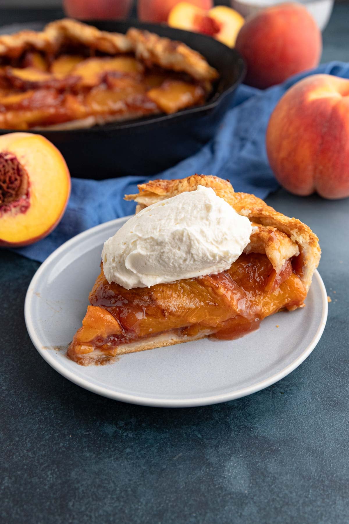A slice of the peach galette with a dollop of whipped cream on a small plate with the rest of the peach galette and fresh peaches in the background.