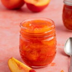 An 8-ounce open jar of homemade peach jam with a small silver spoon next to the jar. There is a another jar of peach jam in the background. There are fresh peaches next to the open jar and in the background.