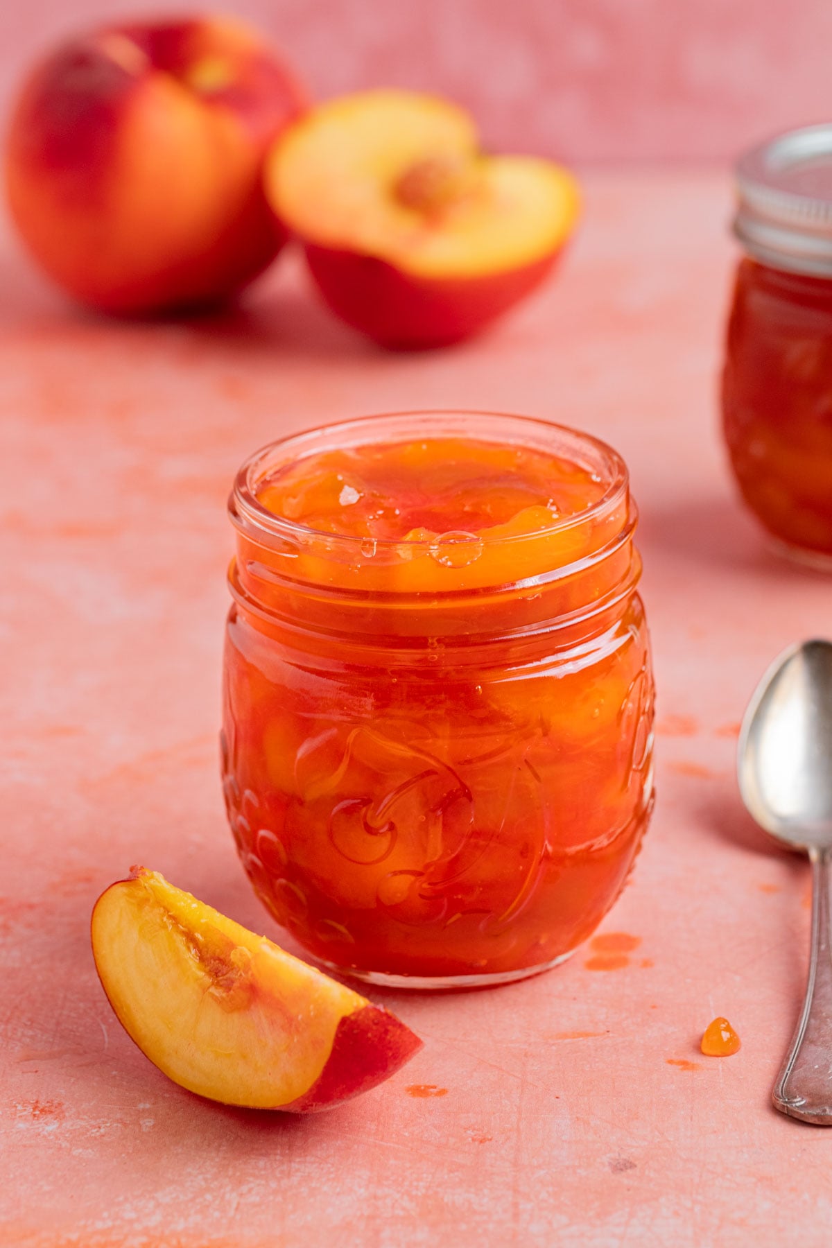 An 8-ounce open jar of homemade peach jam with a small silver spoon next to the jar. There is a another jar of peach jam in the background. There are fresh peaches next to the open jar and in the background.
