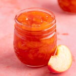 An 8-ounce open jar of homemade peach jam with a small silver spoon next to the jar. There is a another jar of peach jam in the background. There are fresh peaches next to the open jar and in the background.