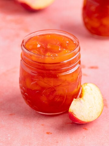 An 8-ounce open jar of homemade peach jam with a small silver spoon next to the jar. There is a another jar of peach jam in the background. There are fresh peaches next to the open jar and in the background.