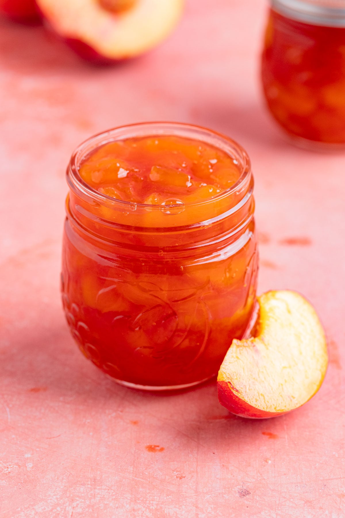 An 8-ounce open jar of homemade peach jam with a small silver spoon next to the jar. There is a another jar of peach jam in the background. There are fresh peaches next to the open jar and in the background.