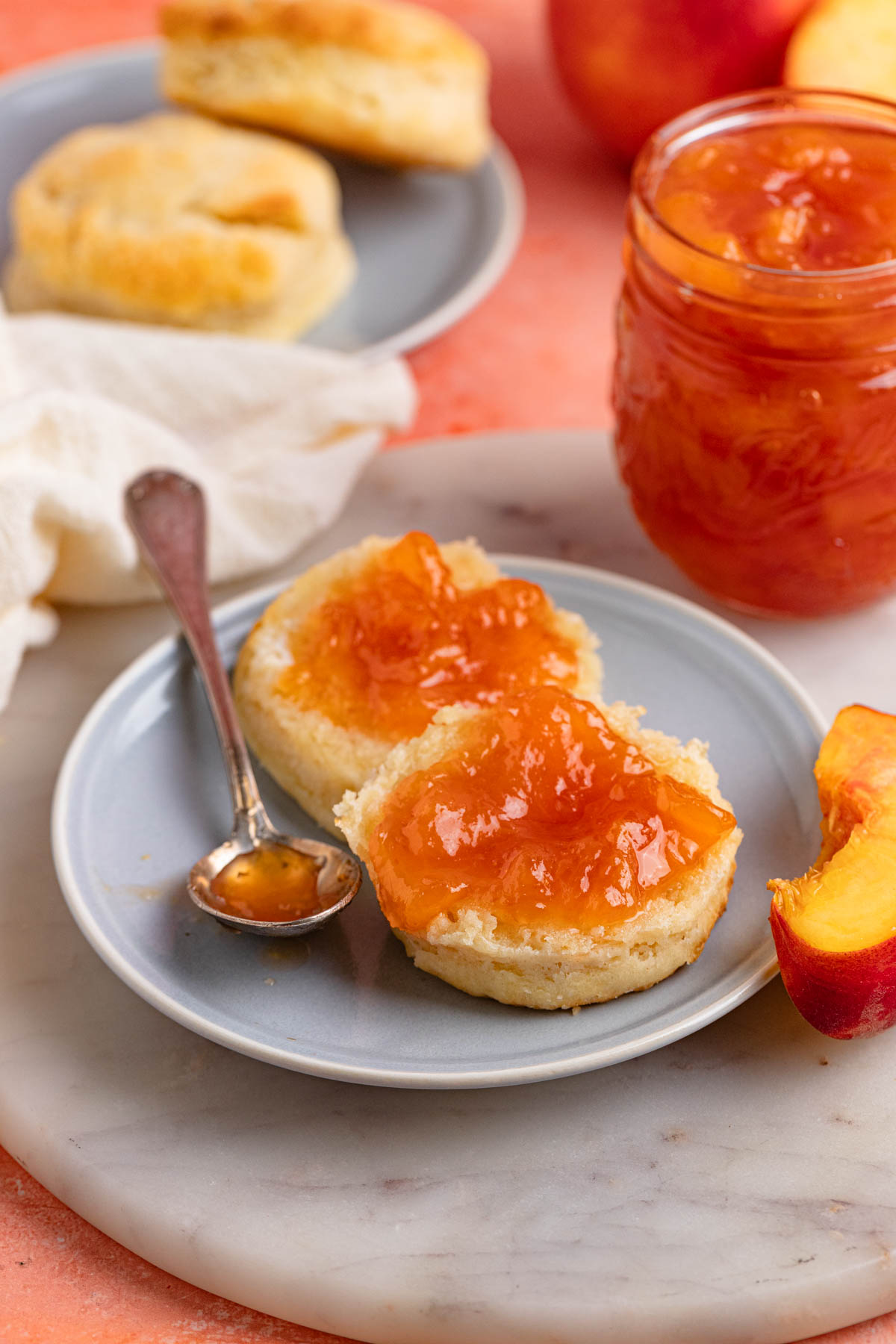A small plate with a biscuit cut in half and peach jam spread on each half. A jar of peach jam in is behind the plate and there is a another small plate with two biscuits to the left and behind the main plate.