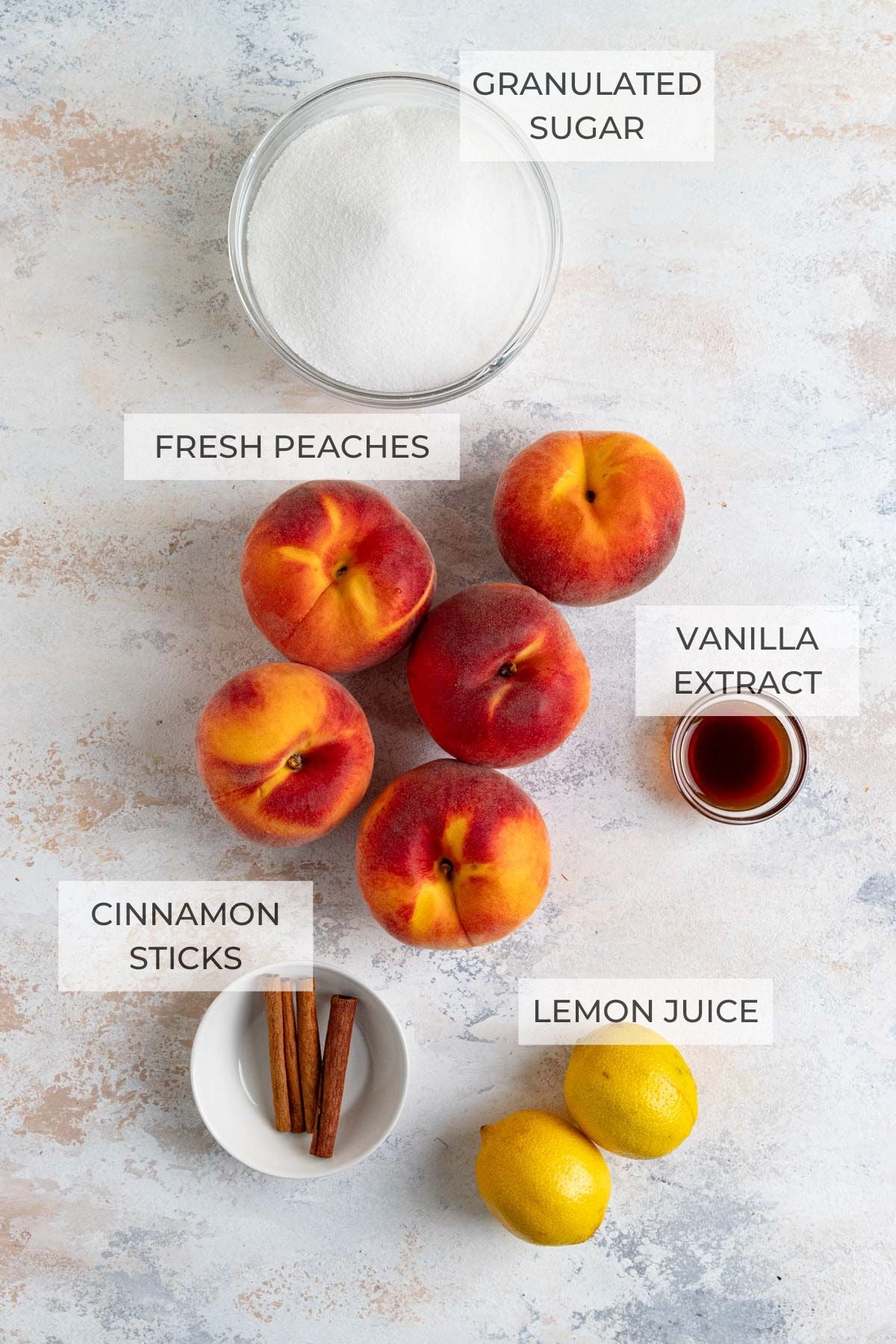 Peach jam ingredients laid out on a board - granulated sugar, fresh peaches, vanilla extract, lemons, cinnamon sticks