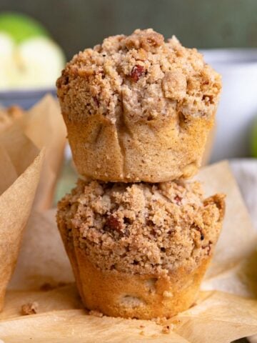 Two apple streusel muffins stacked on top of each other on a wooden board with fresh apples in the background.