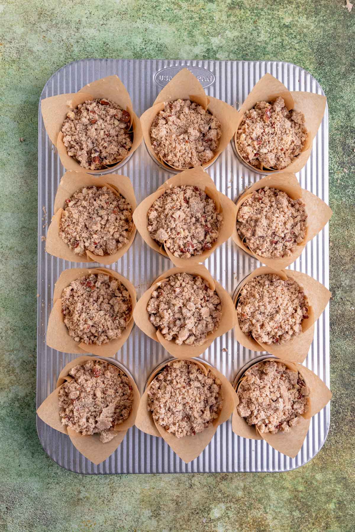 Muffin batter divided between 12 tulip cupcake liners in an aluminum muffin pan. Each muffin is stopped with pecan streusel