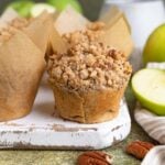 An apple streusel muffin on a white wooden board next to other muffins still wrapped in the muffin liners. There are fresh Granny Smith apples in the background