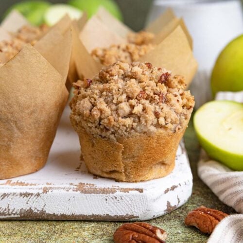 An apple streusel muffin on a white wooden board next to other muffins still wrapped in the muffin liners. There are fresh Granny Smith apples in the background