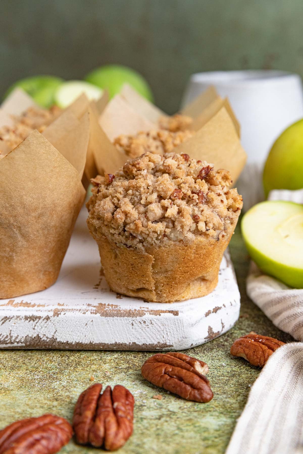 An apple streusel muffin on a white wooden board next to other muffins still wrapped in the muffin liners. There are fresh Granny Smith apples in the background